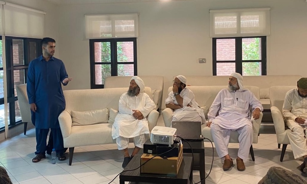 Adil Ahsan standing and speaking to a group of imams seated on sofas, a projector on the table in front of them