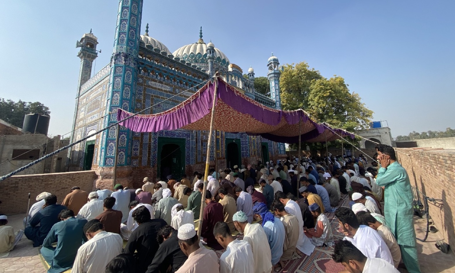 A large congregation seated under an awning outside a mosque faced in turquoise tilework