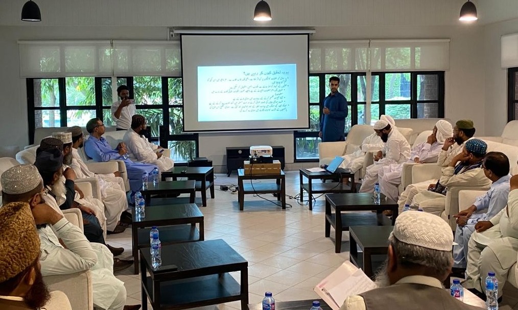 Adil Ahsan presenting beside a projector screen of Urdu slides to imams seated in armchairs around low tables