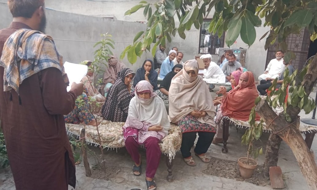 A field team member reading from a sheet of paper to women seated on charpoys in a courtyard, with men seated behind them under a tree